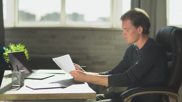 Young thoughtful businessman sitting in the office at the table near notebook and working at documents, make amendment in papers.