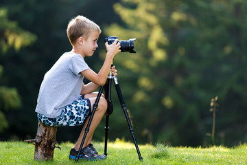 Young blond child boy sitting on tree stump on grassy clearing taking picture with tripod camera.