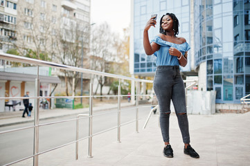 Attractive african american woman with dreads in jeans wear posed near railings against modern multistory building making selfie at mobile phone.