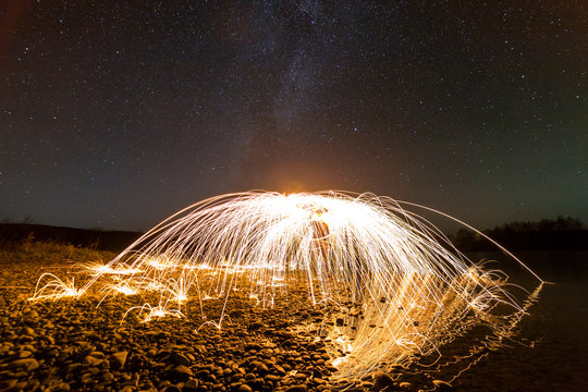Light painting art concept. Spinning steel wool in abstract circle, firework showers of bright yellow glowing sparkles in fountain form on river bank on blue night starry sky copy space background.