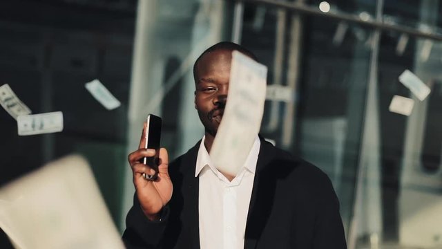 Portrait of african american businessman looking at camera while money flying down all over the place. He holding smartphone in hands. Money rain on the businessman in slow motion