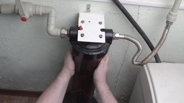 Plumber unscrews glass flask with old water filter in basement.
