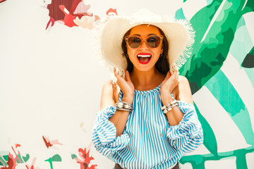 Beautiful lady in a striped T-shirt and denim shorts, sunglasses and a straw hat. It stands on the background with a tropical pattern. concept of summer holidays and shopping