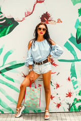 Beautiful lady in a striped T-shirt and denim shorts, sunglasses and a straw hat. It stands on the background with a tropical pattern. concept of summer holidays and shopping