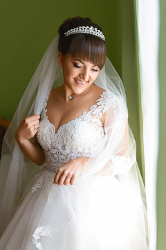fashion photo of beautiful bride with dark hair in elegant wedding dress and diadem posing in room in the wedding morning