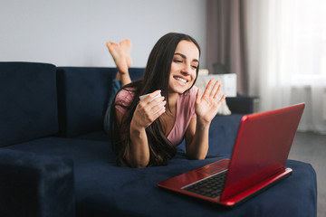 Attractive young woman lyong on sofa. She waves with hand and smile. Model look on laptop screen. She hold white cup.