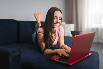 Attractive beautiful young woman lying on sofa in room. She type on laptop keybaord and smile. Model pose. She looks happy.