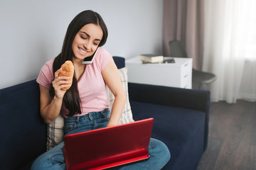 Cheerful young woman work at home. She sit on sofa and type on laptop keyboard. Model talk on phone and hold croissan in hand.