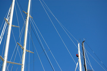 Masts of sailing boats, with a spreader, shrouds. A sailor is climbing up on a mast. A blue sky in the background.