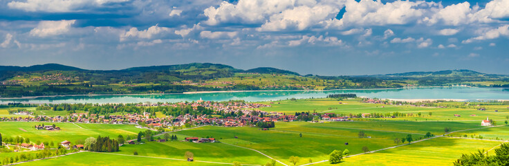 A beautiful landscape panorama photo of the village Schwangau with its green fields, the turquoise blue Forggensee lake and the natural surrounding in the district of Ostallgäu in Bavaria, Germany.