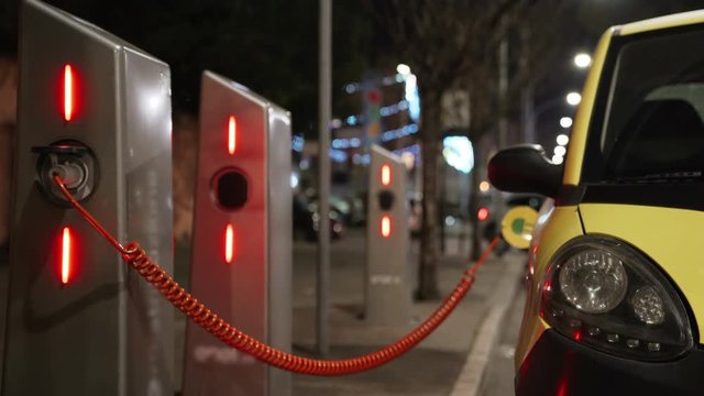 Small electric car at charging station in the city street on the background of driving cars in the night. Eco transport and power supply. 4K.