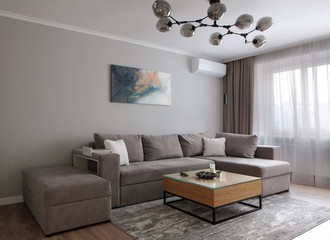 Grey corner couch with three pillows standing in bright living room interior with painting and carpet.Lightning off