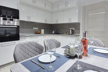 White kitchen interior with grey chairs and served table with white plates glasses and moss on the wall in new luxury home with lights on.
