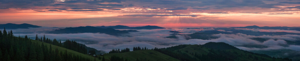 Naklejka na ściany i meble Panoramic photo of Mountain range through the morning colorful fog in the summer. Beautiful sunrise in the Carpathian Mountains with visible silhouettes and pink clouds in the sun rays.