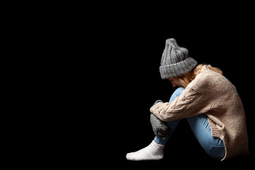 The girl is sitting alone on the floor on a black background of emptiness, hugging her legs with...