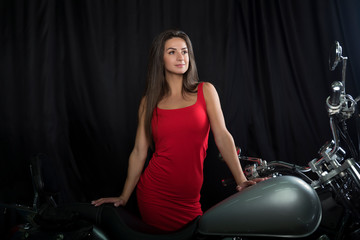 Fototapeta na wymiar A beautiful young brunette in a bright red dress stands near a chrome motorcycle on a black background