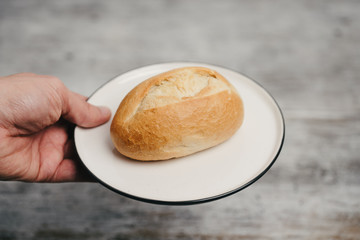 Man's hand holding plate with German style bread roll or bun, so called Schrippe or Brötchen, a small loaf made of wheat flour traditionally served for breakfast on white plate and wooden background