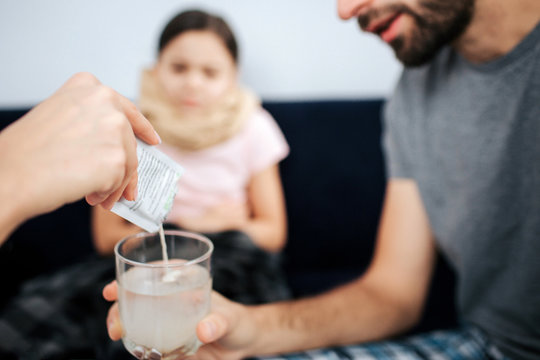 Cut view of doctor's hand pouring powder medicine into glass of water. Young man hld it. Small sick girl sit on sofa and look at process.