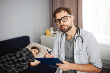 Serious young male doctor look on camera through glasses. His ptient lying on bed. Small girl covered with blanket. She sick.