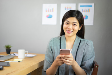 Smiling Business girl working in office with laptop,Happy Asian business woman using phone sitting on a chair at modern home studio.Concept of young people working mobile devices,contact to customer
