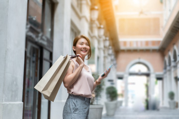 Smiling Asian Thai girl holding gift in shopping bags She Compares prices on mobile smartphones looking for clothes in a dress shop  - Concepts of relaxing lifestyle,shopping online technology in city