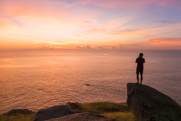 Fototapeta premium silhouette of young man stands on the stone and takes a photo with small boat and beautiful amazing sea sunset of orange red colors
