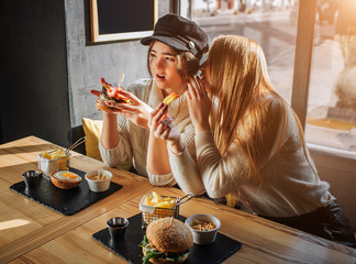 Beautiful young women sit in side at table. They gossip. Blodne girl wisper on ear to another girl....