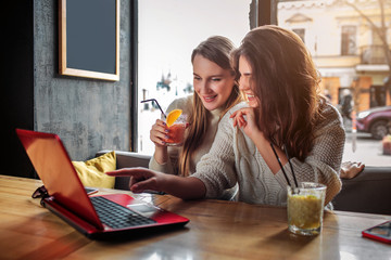 Beautiful and positive young women watch movie on laptop. They smile. Model hold glass of drink. Another points on screen.