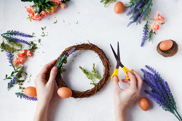 Female hands make Easter wreath of flowers, herbs and eggs on a light background. Easter concept