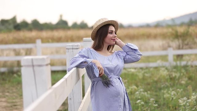 Pregnant woman in nature try on knitted hat. Background of field and white fance near the farm