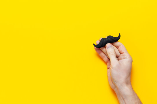 Men's hands hold Black mustache, props for photo booths, carnival, parties on yellow background top view flat lay copy space Father's day Creative party decoration concept