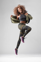 Happy beautiful African American Girl with wild curly hair and wearing an green sports fitness wear in studio with gray background doing a graceful jump