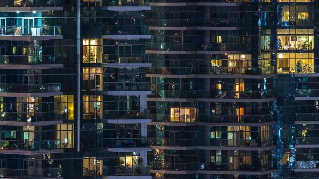 Glowing windows in multistory modern glass residential building light up at night timelapse.