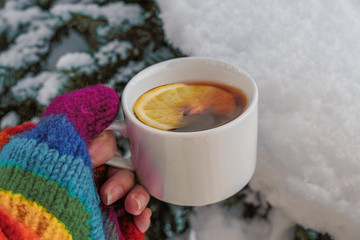 hand in a cozy multi colored woolen glove holding a cup of hot tea with a lemon in the winter forest against the background of white snow and fir tree branches