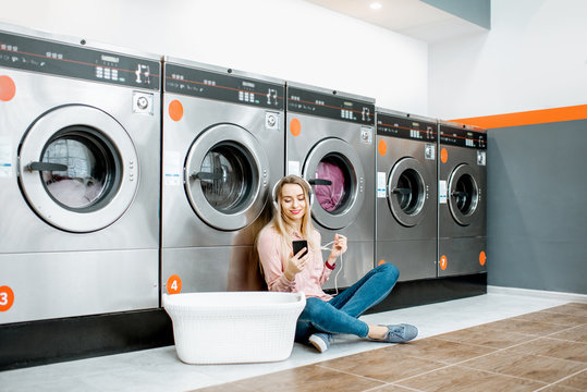 Young woman waiting for the clothes to be washed sitting on the floor and listening to the music at the self-service laundry - Powered by Adobe