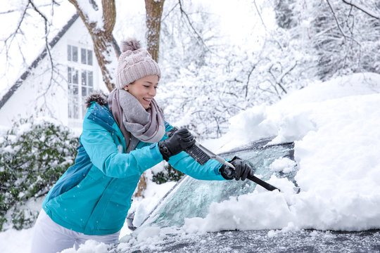 h&uuml;bsche junge blonde frau schippt im winter den wei&szlig;en Schnee vor ihrem haus auf einen grossen haufen - mit k&auml;lteschutz blauer jacke und m&uuml;tze bekleidet l&auml;chelt sie gl&uuml;cklich