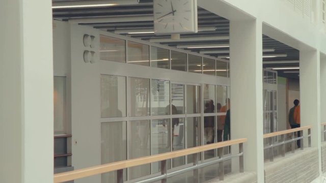 People walk down a bright interior hallway past a room with a glass wall. Modern architecture, sunlight and white paint make a bright scene. A clock hangs over a hallway and railing.