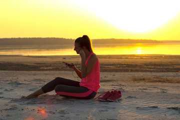 Young beautiful girl in sportswear sits on a sandy beach and enjoys an orange sunset by the sea and listens to music in her mobile phone