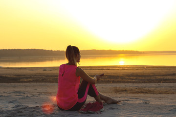 Fototapeta na wymiar Young beautiful girl in sportswear sits on a sandy beach and enjoys an orange sunset by the sea and listens to music in her mobile phone