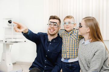 Happy young family at ophthalmologist appointment. Dad denim shirt dark hair smiles, shows index...