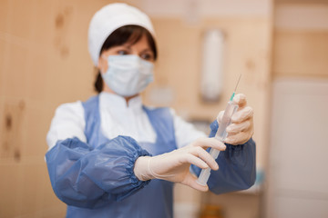 young nurse in special sterile clothing in a protective mask is standing indoors holding a syringe...