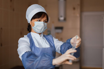 Nurse in a white mask in blue medical clothing stands holding a syringe in her hand. A female...