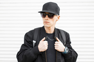 Portrait of an attractive man hipster in a black cap in stylish black sunglasses in a trendy jacket...