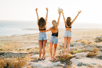 Back view. Back of three girls holding hands up wearing hat and jeans enjoying the beach views...