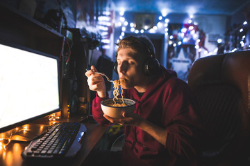 Young man eats noodle soup with his eyes closed and uses a computer at home in the evening. Portrait of a gamer eating a fast cooking soup at the computer table and playing video games