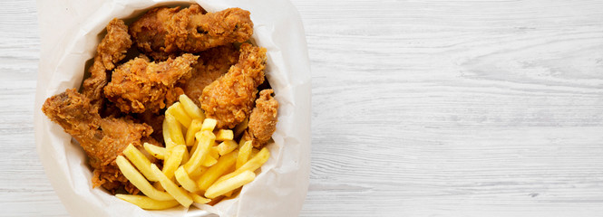 Fast food: fried chicken drumsticks, spicy wings, French fries and tender strips in paper box over...