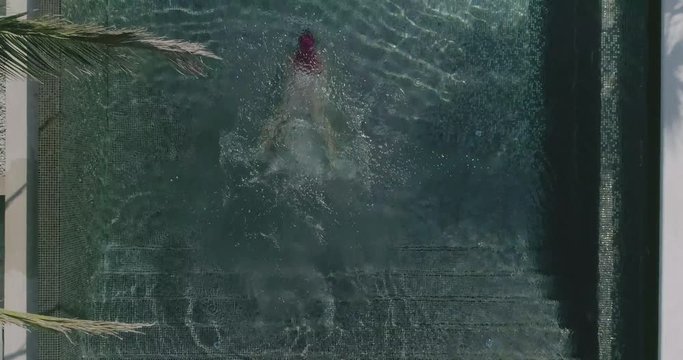 AERIAL CLOSE UP TOP DOWN: Young woman on summer vacation jumping head first into clear blue pool water for refreshment on hot sunny day. Girl in pink bikini diving into empty pool in luxury resort
