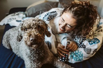 .Young and funny woman playing with her nice spanish water dog on top of the bed, freshly awake on...