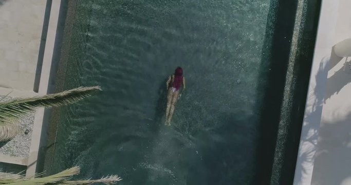 AERIAL CLOSE UP TOP DOWN: Young woman on summer vacation jumping head first into clear blue pool water for refreshment on hot sunny day. Girl in pink bikini diving into empty pool in luxury resort