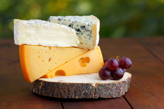 Camembert, brie, hard cheese and blue cheese on wooden board. Slices of various cheeses and grapes on blurred background. Dorblu and soft cheese on wooden boards. Cheese and grapes on bokeh
